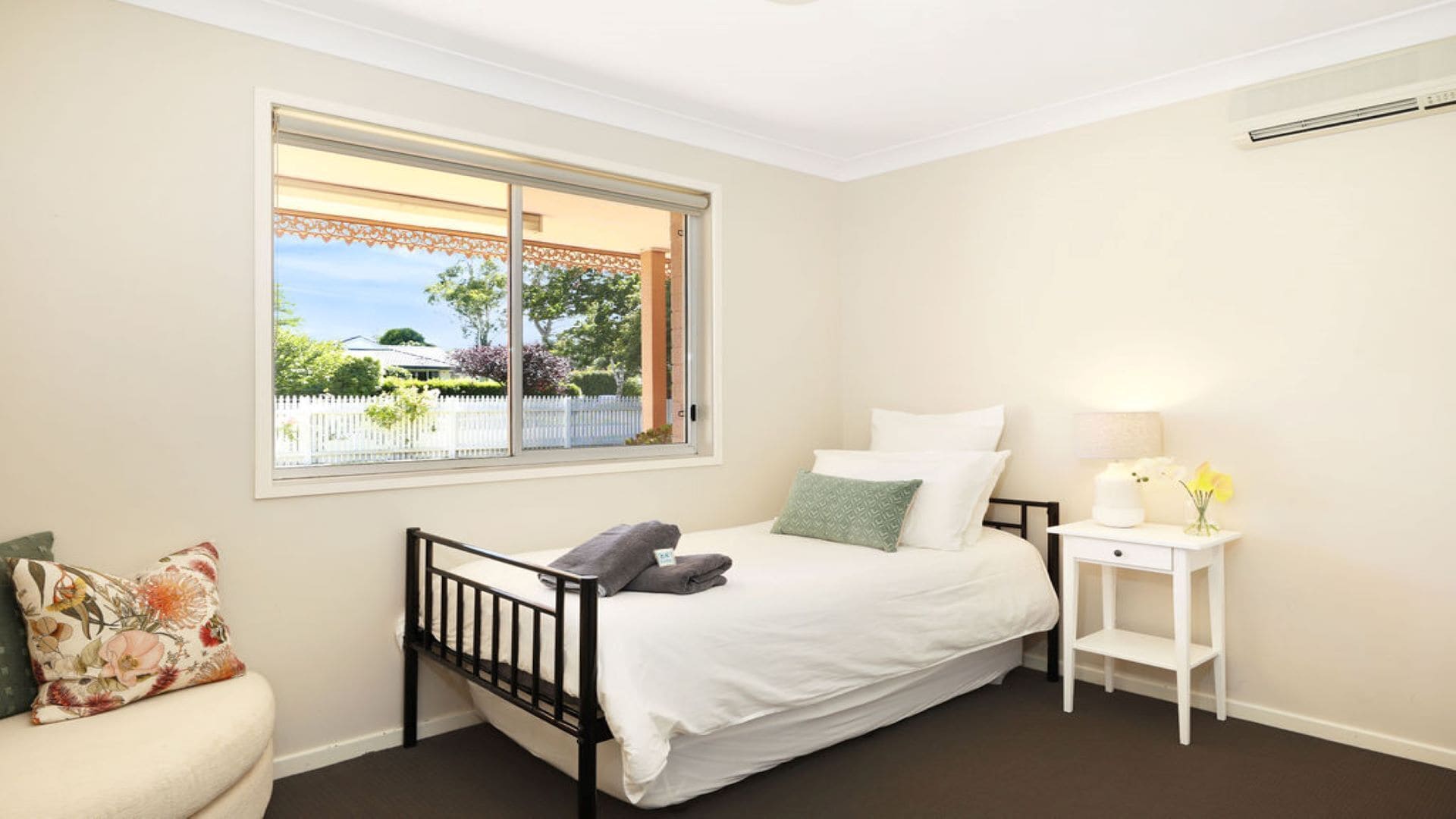 A bedroom with a bed, a nightstand, a chair, and a window with a view of a fence and greenery.