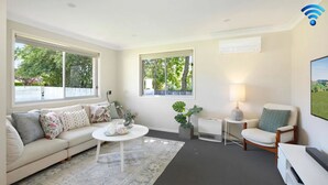 A living room with a sofa, coffee table, and a chair. There are two windows with views of greenery. - That Pink House! – pets welcome (Bowral)