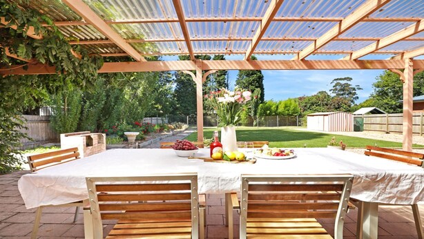 An outdoor patio with a wooden pergola, a table set with fruits and flowers, and a bench. - That Pink House! – pets welcome (Bowral)