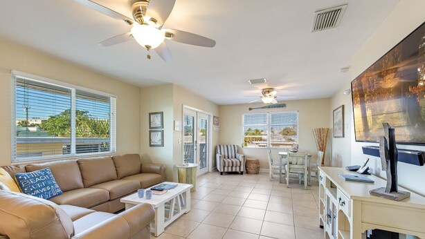 A spacious living room with a beige leather sofa, a dining table with chairs, a ceiling fan, and a large window with blinds. - Island Charm with exceptional private pool is ready to rent (Fort Myers Beach)