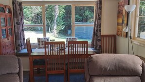 A dining area with a table and chairs, a wooden cabinet with glass doors, and a view of the garden through large windows.