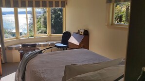 A room with a bed, a desk, a chair, and a window with a view of the sea.