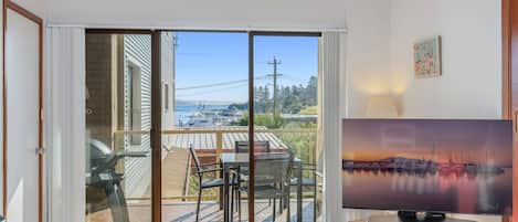 A room with a sliding glass door leading to a balcony with outdoor furniture and a view of boats and hills.