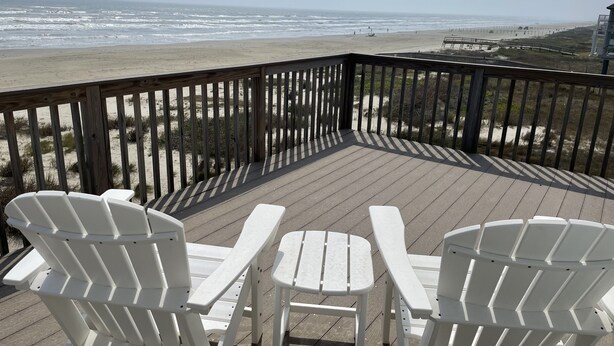 A wooden deck with white Adirondack chairs and a view of the beach.