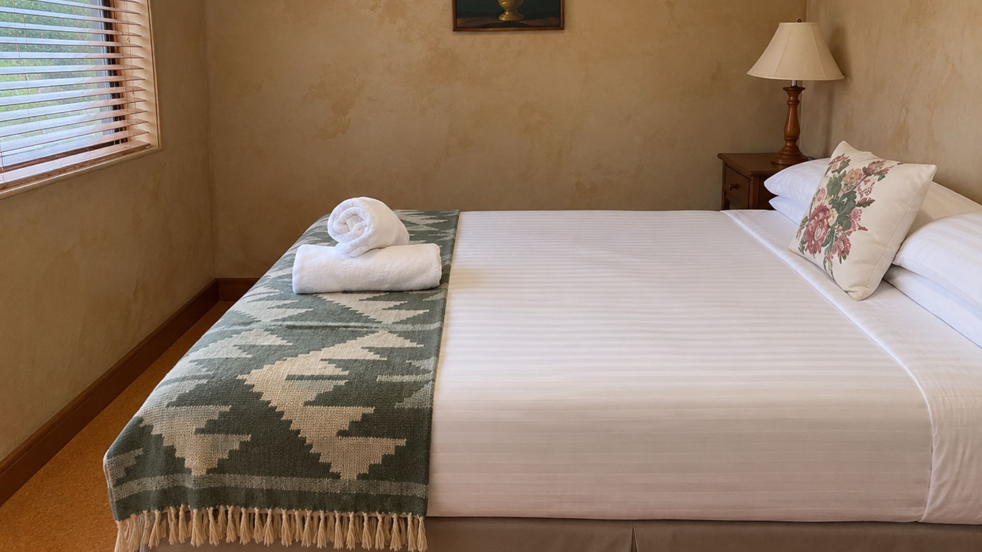 Een hotelkamer met een bed, een raam met luxaflex, een nachtkastje met een lamp en een aan de muur hangend schilderij.