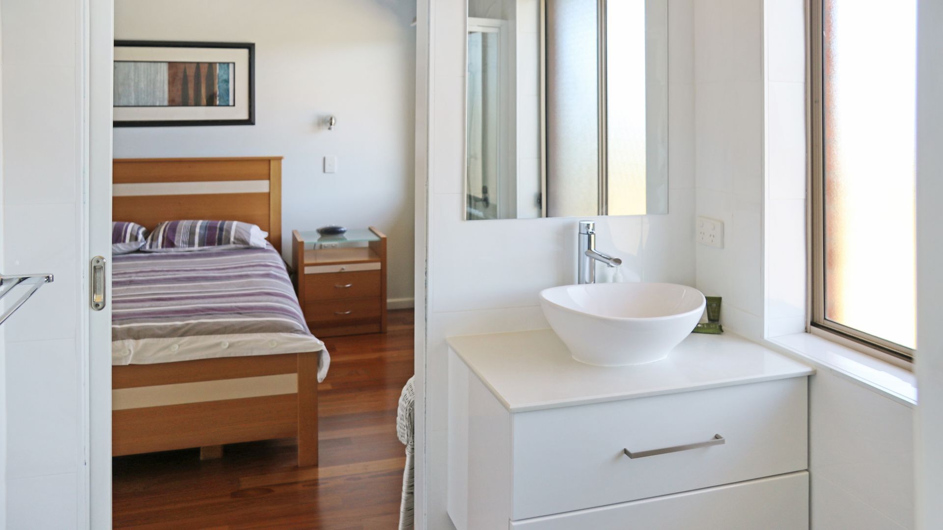A modern bedroom with a wooden bed, a white bedside table, a large mirror, and a white bathroom with a vessel sink.