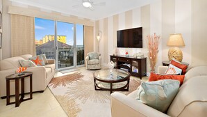 A spacious living room with a large flat-screen TV, comfortable seating, a coffee table, and a view of buildings through large glass doors.
