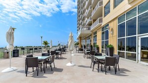 An outdoor dining area with tables and chairs, umbrellas, and a building with balconies and large windows.