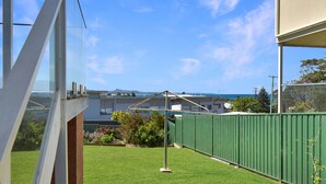 A balcony with a view of the sea and mountains, a green fence, and a grassy area with plants.