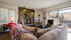 Un salon chaleureux avec une cheminée en pierre, un téléviseur et un coin salon confortable offrant une vue sur l’extérieur.