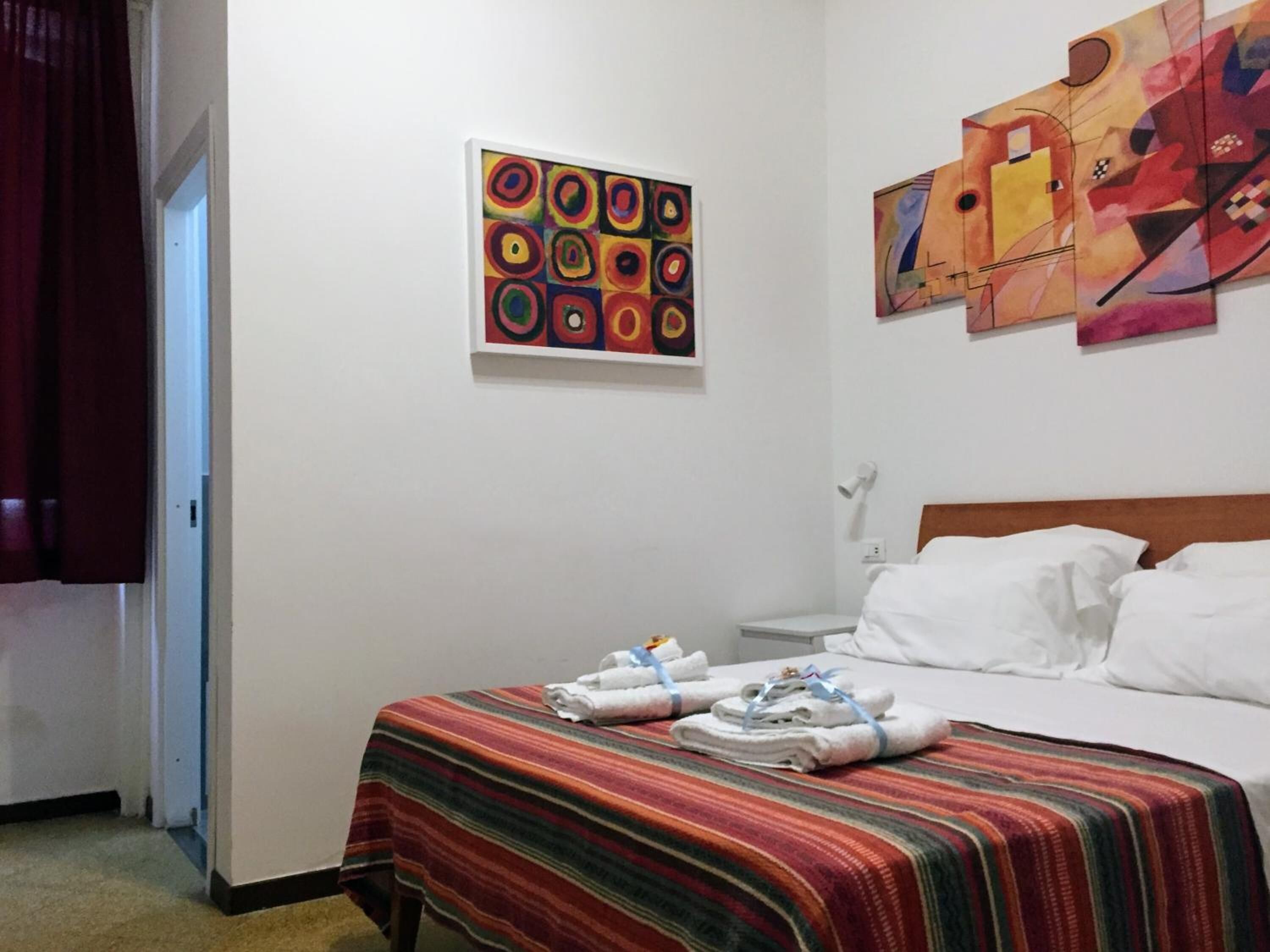 Une chambre d’hôtel avec un lit, une table de chevet, une lampe, un mur orné de deux œuvres d’art et une couverture à rayures posée sur le sol.