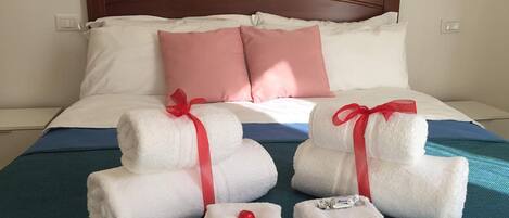 A neatly made bed with white and pink pillows, a teal bedspread, and three white towels with red ribbons tied around them.