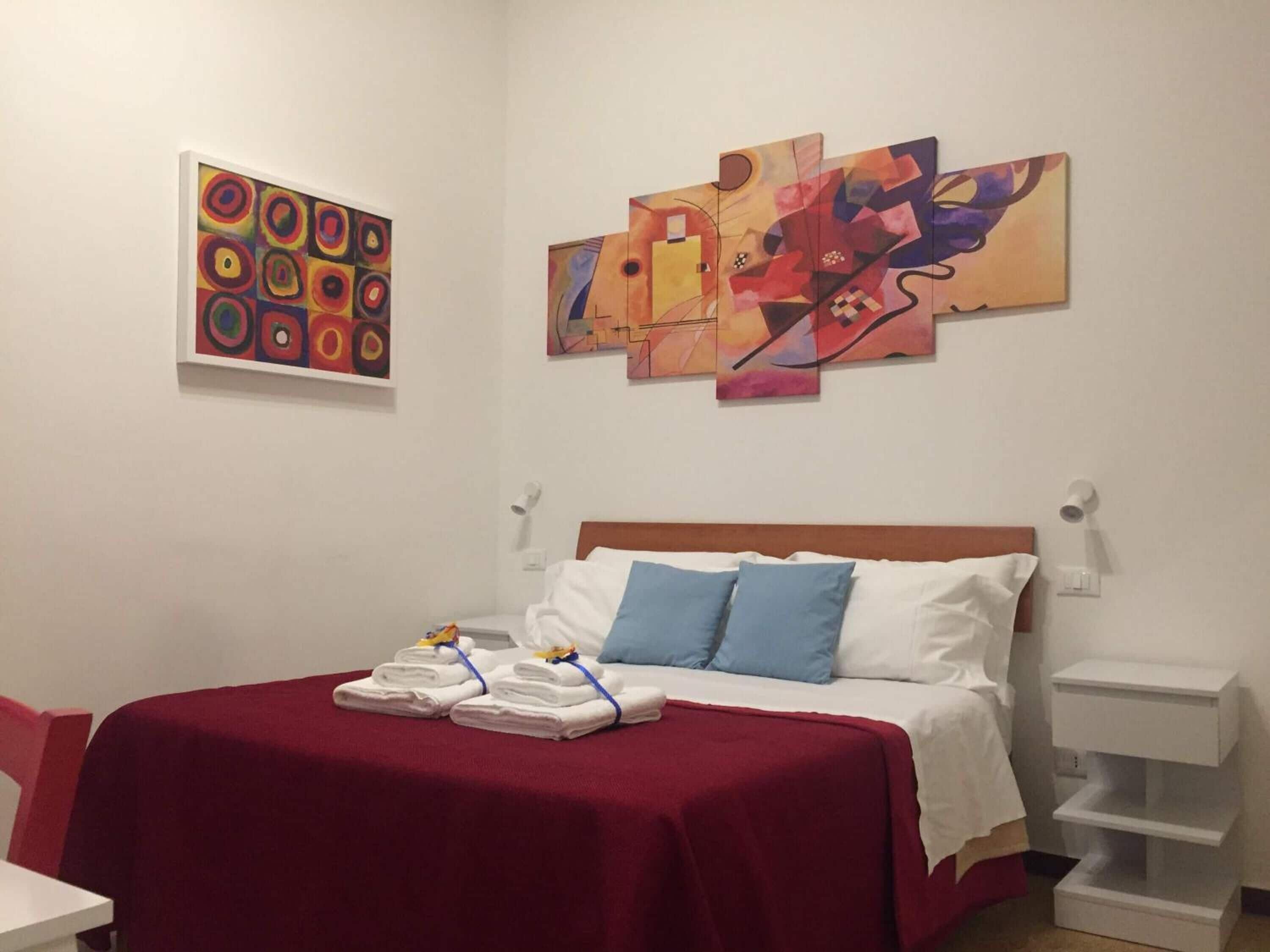 Una camera d'albergo con un letto, una coperta rossa, cuscini bianchi e opere d'arte alle pareti.