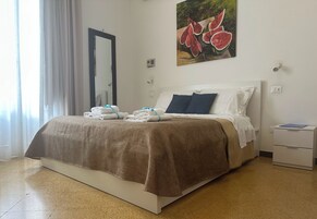 Un letto rifatto con una coperta marrone, cuscini bianchi e un cuscino decorativo blu. Un comodino bianco con una cartella blu sopra. Un quadro con angurie appeso al muro.