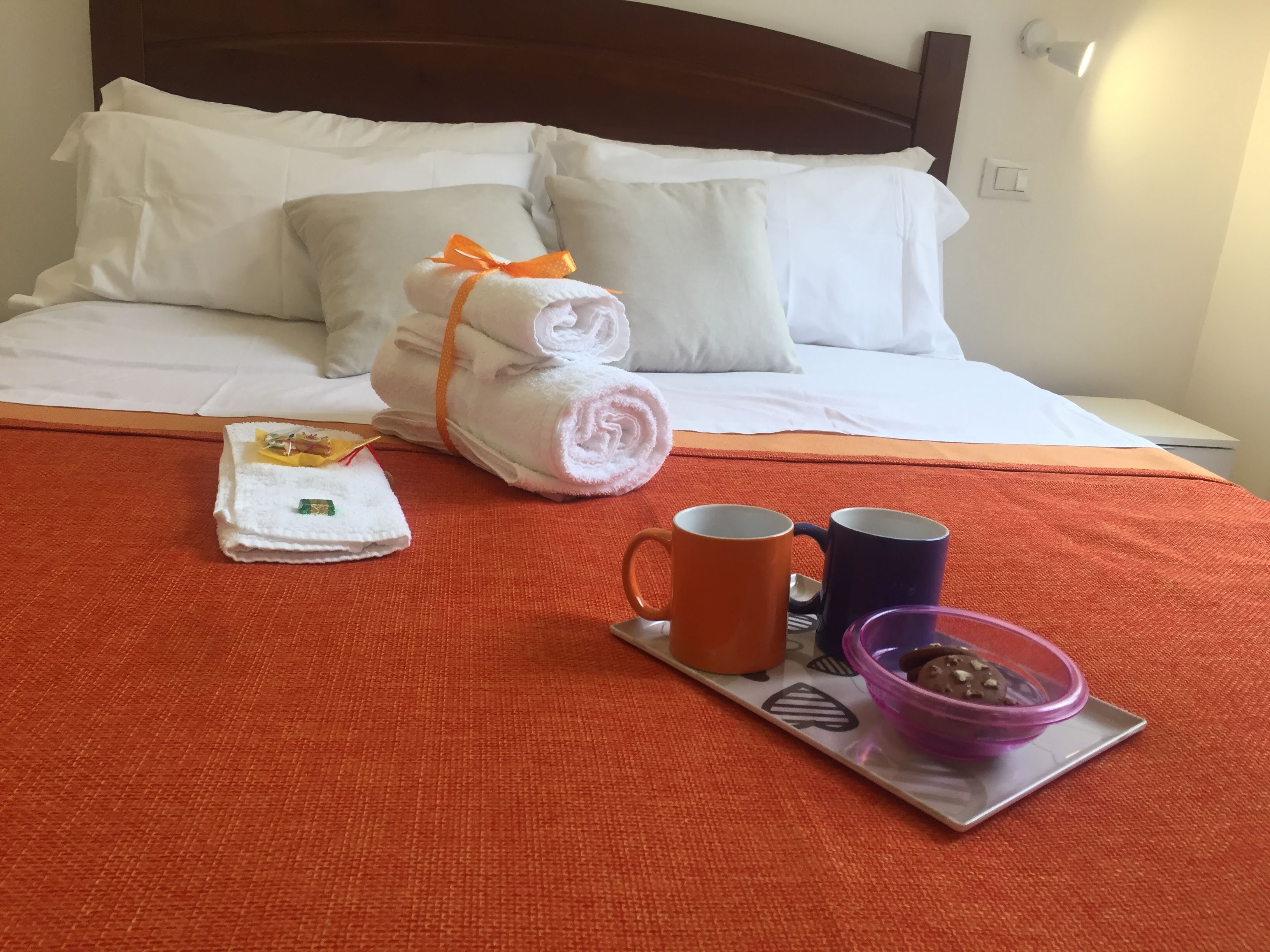 A neatly made bed with white linens and pillows, a tray with a mug, a bowl, and a folded towel with an orange ribbon.
