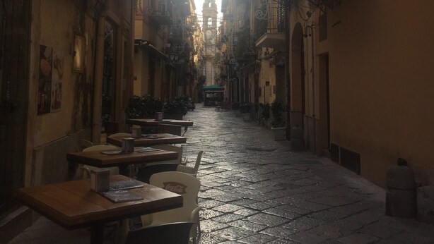 A narrow street with buildings on both sides, outdoor seating, and a clock tower in the distance. - Yooroom (Palermo)