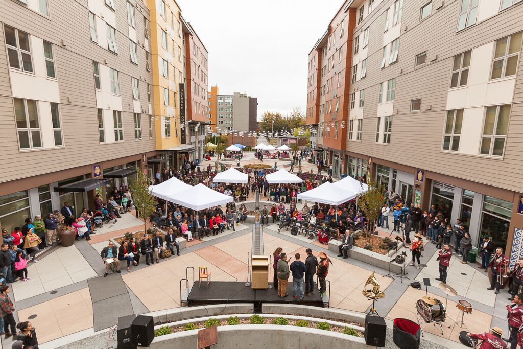 A courtyard with a central stage, surrounded by seating and covered areas, in a multi-story building with commercial spaces on the ground floor.