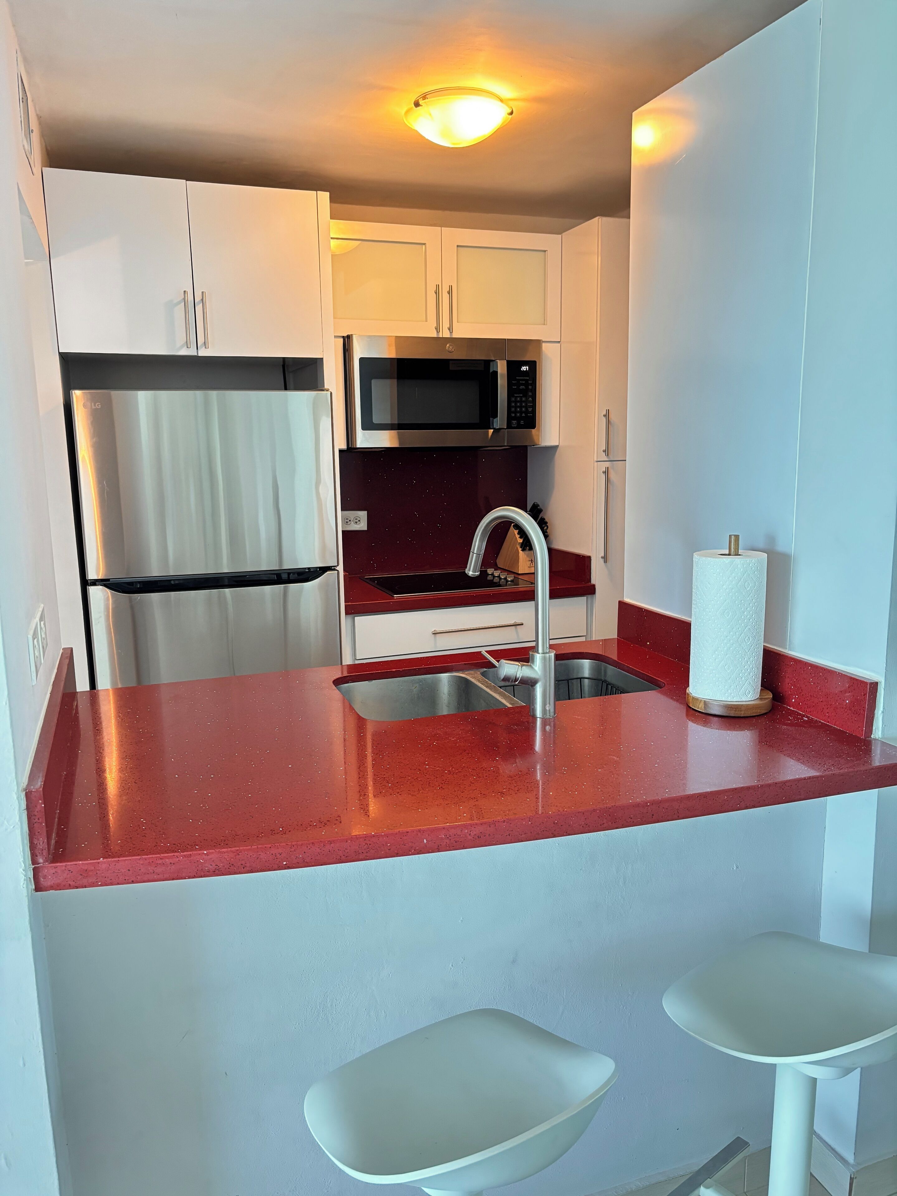 Cocina compacta con armarios blancos, refrigerador de acero inoxidable, microondas, lavamanos con grifo y encimera roja.