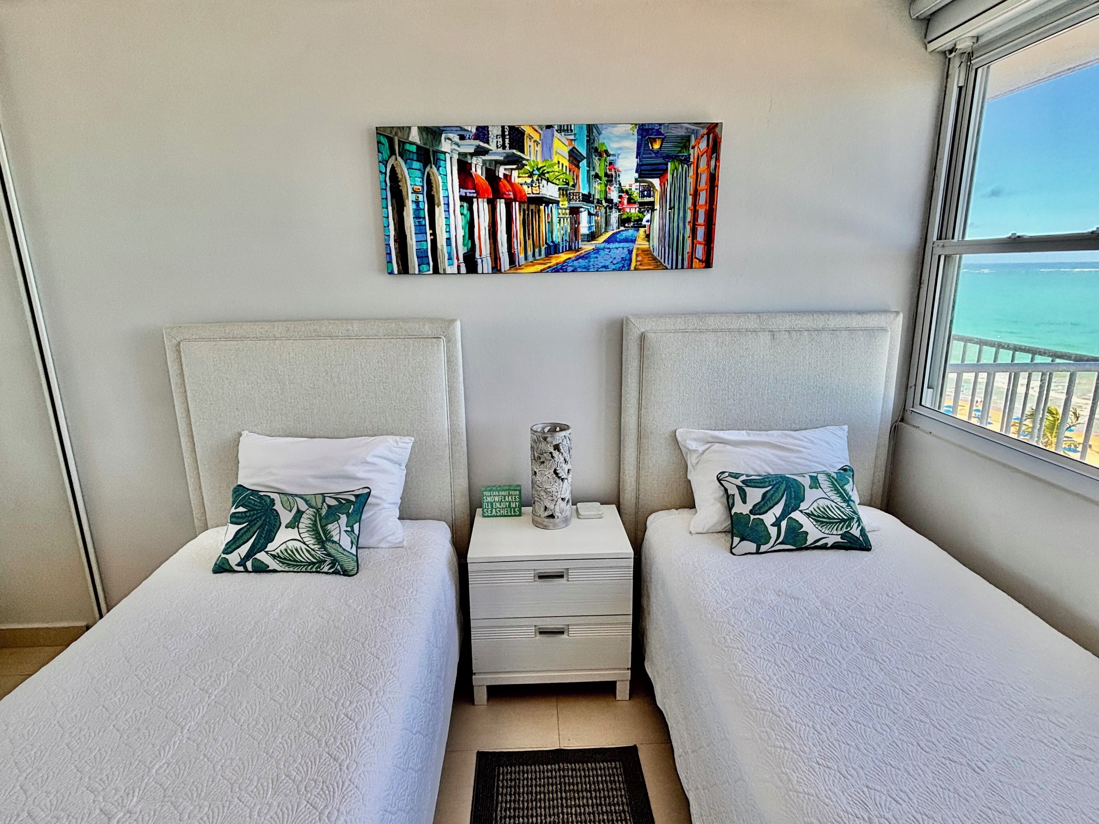 A hotel room with two beds, a nightstand, a painting, and a window with a view of the sea.