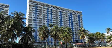 A high-rise hotel with multiple balconies, palm trees, and a sandy ground area.