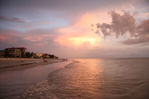 Ein Strand mit Gebäuden im Hintergrund und einem Sonnenuntergang über dem Wasser.