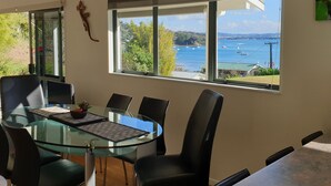 Uma zona de refeições moderna com mesa de vidro e cadeiras pretas, com vista para uma baía cénica com barcos.