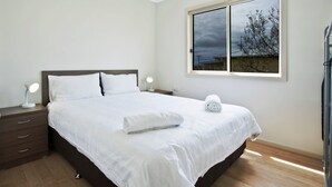 Un lit bien fait, avec du linge de lit blanc, une lampe de chevet et une fenêtre donnant sur des arbres et un ciel nuageux.