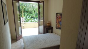 Een slaapkamer met een bed, een klein tafeltje met een lamp, een schilderij aan de muur en uitzicht op groen door een open deur.
