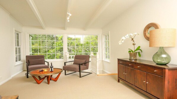 A room with two chairs, a small table, a cabinet, and a large window with a view of greenery. - Que Sera – pet friendly (Bowral)
