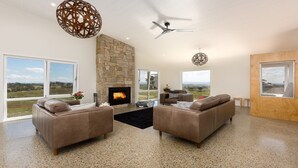 A spacious living room with a stone fireplace, leather sofas, a ceiling fan, and large windows offering a view of the outdoors.