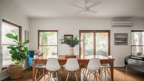 Ein Essbereich mit Holztisch und weißen Stühlen, großen Fenstern mit Blick auf Bäume, einem Deckenventilator und einer Klimaanlage.