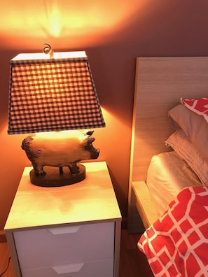 Cozy bedroom with a bedside table, lamp, and wooden sculpture, featuring a patterned blanket and simple headboard.
