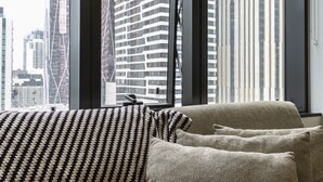A modern living room with a sofa and pillows, large windows with blinds, and a cityscape view.