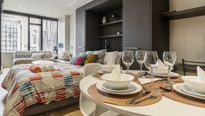 A modern hotel room with a neatly made bed, a dining table set for two, and a large window offering a city view.