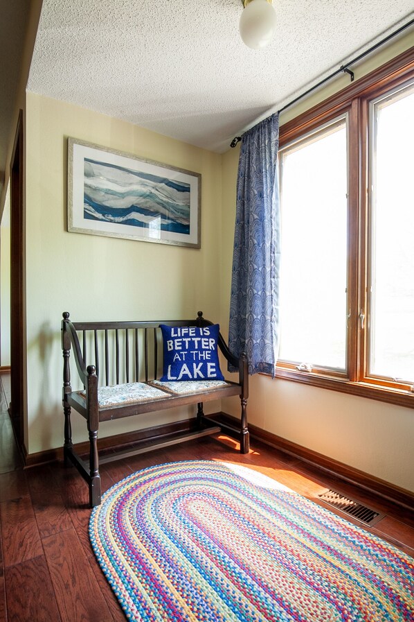 Una habitación con un banco de madera, un tapete tejido y colorido, un cuadro en la pared y una ventana con cortina.