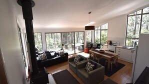 A modern living room with a fireplace, a sofa, a dining table, and a kitchen area with a stove and sink. - 11 Burrawong Place - Bushland Haven (BARRAGGA BAY)