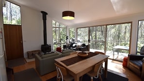 A cozy living room with a fireplace, a dining area, and large windows offering a view of the forest. - 11 Burrawong Place - Bushland Haven (BARRAGGA BAY)