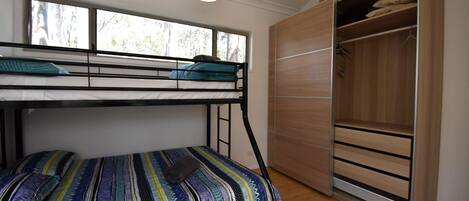 A bunk bed with a patterned bedspread, a wooden wardrobe, and a window with a view of trees.