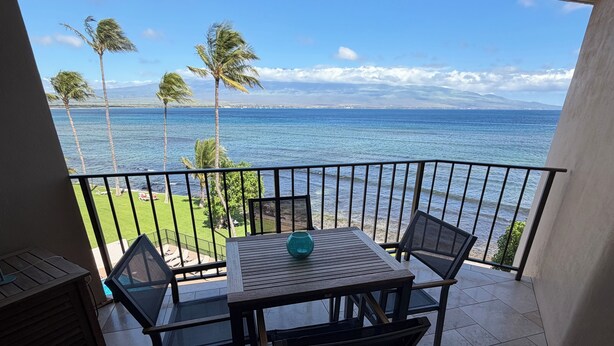 Varanda com mesa e cadeiras, vista para uma praia com palmeiras e o oceano.
