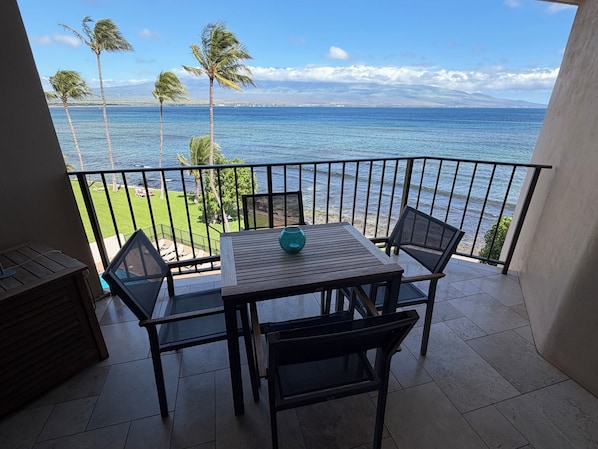 A balcony with a table and chairs, overlooking a beach with palm trees and the ocean.