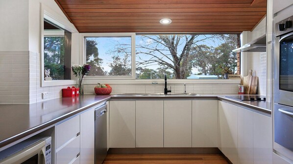 A modern kitchen with white cabinetry, a wooden ceiling, and a window overlooking trees.
