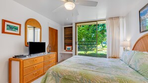 Ein Schlafzimmer mit Bett, Fernseher auf einer Kommode, Deckenventilator und einem Fenster mit Blick auf Grünflächen.