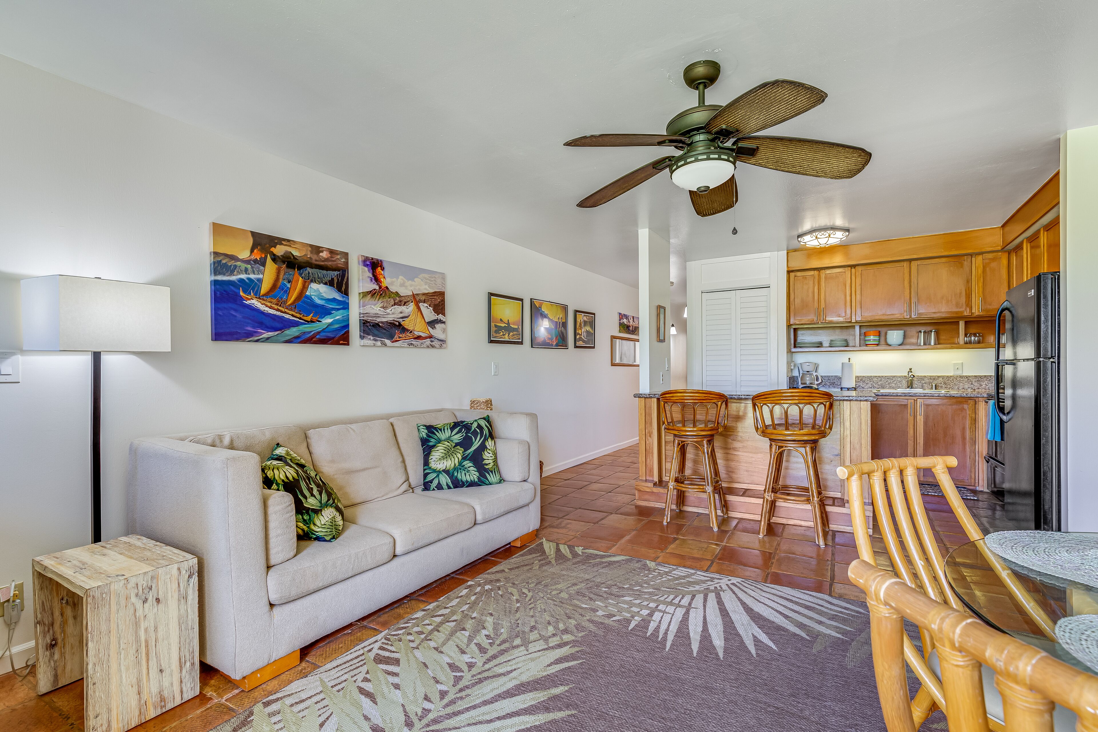 A modern living room with a sofa, a coffee table, and a dining area with chairs. There is a kitchen area with wooden cabinets and a refrigerator.