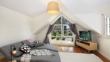 A cozy loft bedroom with a large window, a gray sofa, a TV, and a wooden cabinet.
