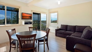 A modern living room with a round dining table, wooden chairs, a gray sofa, and large windows offering views of the outdoors. - Seaview Beach House 2 (BERMAGUI)