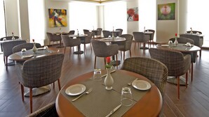 A spacious dining area with multiple round tables, each set with plates, cutlery, and glasses. The room features a high ceiling with decorative lighting fixtures and large windows allowing natural light. - Dunes View Muscat  Hotel Apartments  (Muscat)