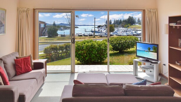 Un salon avec un canapé, un téléviseur et une vue sur une marina, visible à travers une porte vitrée.