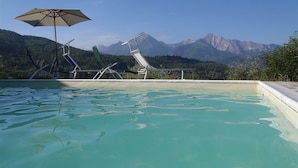 Piscina con sillones de descanso y una sombrilla, rodeada de montañas.