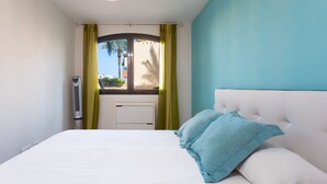 Een slaapkamer met een bed, een raam met gordijnen, een radiator en uitzicht op palmbomen en gebouwen.