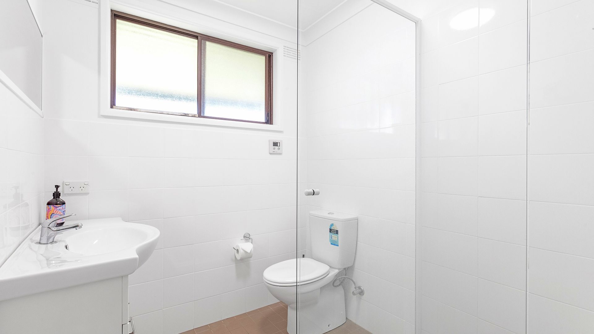 A small, clean bathroom with white tiles, a white sink, a toilet, and a window above the sink.
